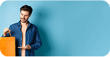 Homem de pele clara, cabelo castanho curto, usando jaqueta jeans azul e blusa rosa, segurando sacola de compras laranja sobre fundo azul