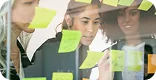 Equipe de três pessoas em sessão de design de experiência do usuário, escrevendo e colando anotações verdes em parede de vidro