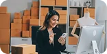 Profissional de e-commerce trabalhando em computador, sorrindo enquanto confere pedidos, caixas de envio empilhadas ao fundo