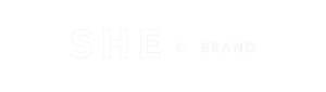 Logotipo da She Brand em branco, com a palavra “SHE” em letras maiúsculas destacadas à esquerda, um ponto central como elemento gráfico e a palavra “BRAND” em letras mais finas à direita, sobre fundo transparente.